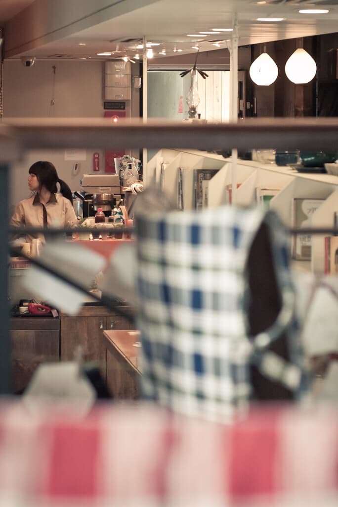 A woman behind a café counter, seen past a blurred checked cloth and shelves in the foreground.
